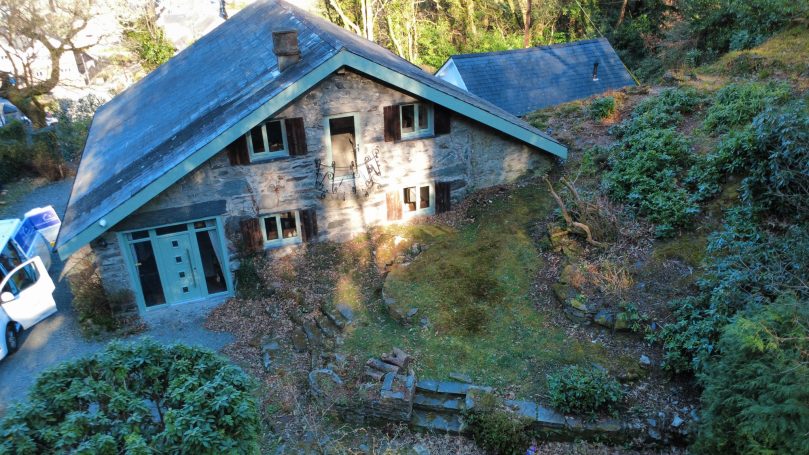 Derelict stone house surrounded by overgrown greenery and trees.