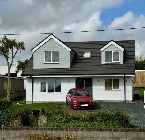 Two-storey house with a grey roof and red car parked in front, surrounded by greenery.